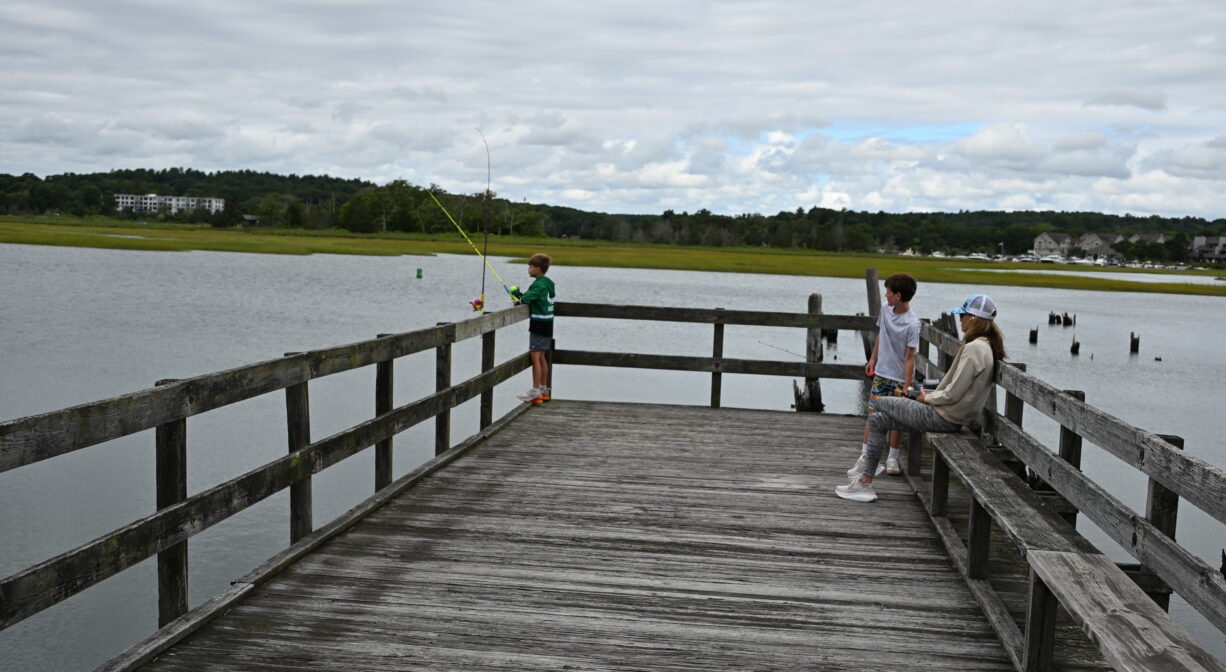 A photograph of a few individuals fishing on a dock beside a river.