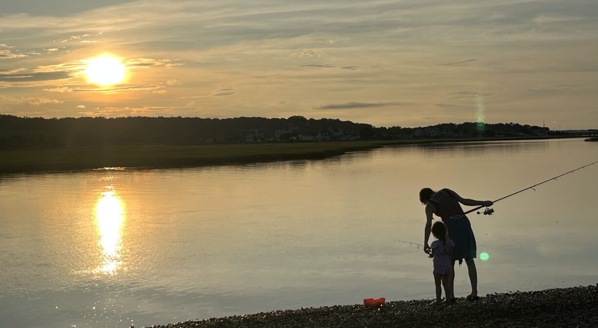 A photograph of an adult and a child at sunset, fishing on a river.