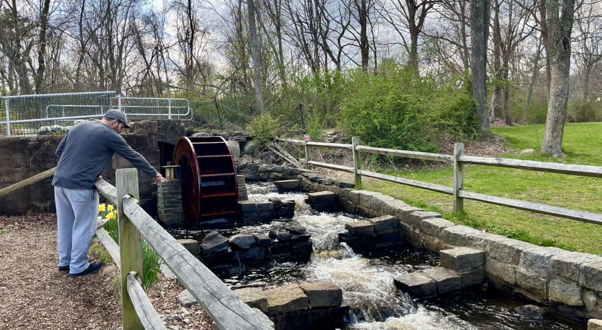 A photograph of an adult pointing into a stream as it flows over a fish ladder in a park.