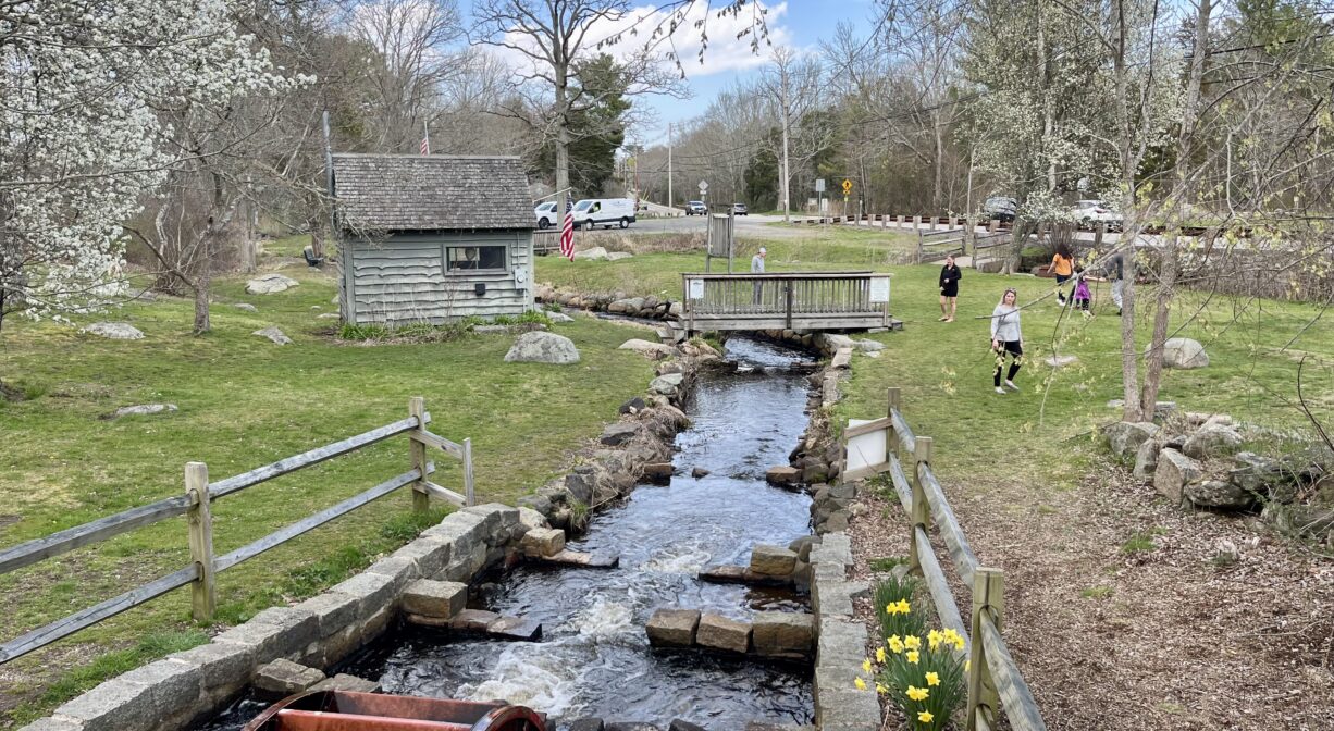 A photograph of a grassy park with a water wheel, a stream flowing down the middle, and a few individuals on the side.