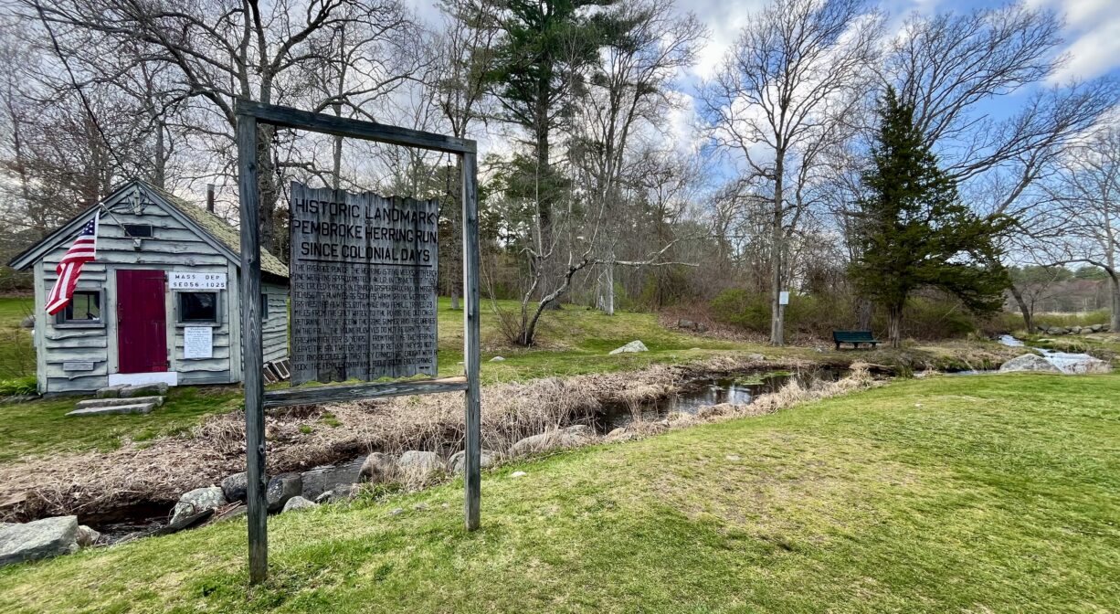 A photograph of a grassy park with a property sign and a shed, and stream flowing through it.