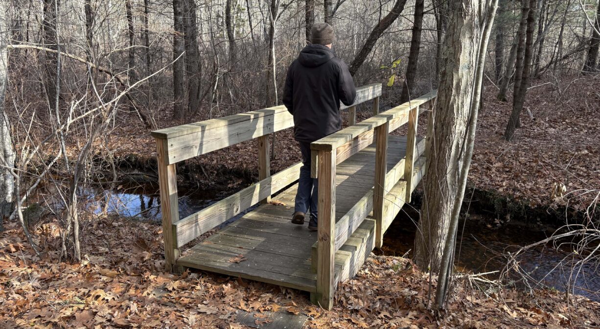 A photograph of a person walking across a wooden bridge in a forest.