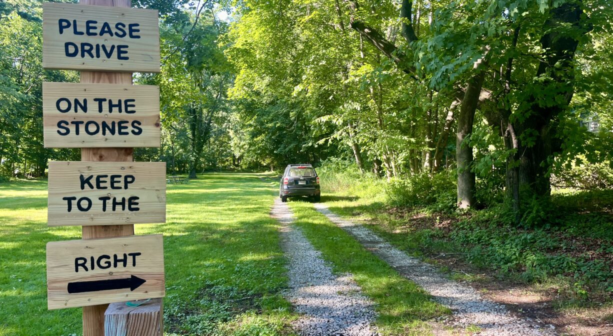 A photograph of an access road to a grassy property bordered with trees, with signs in the foreground and a road to one side.