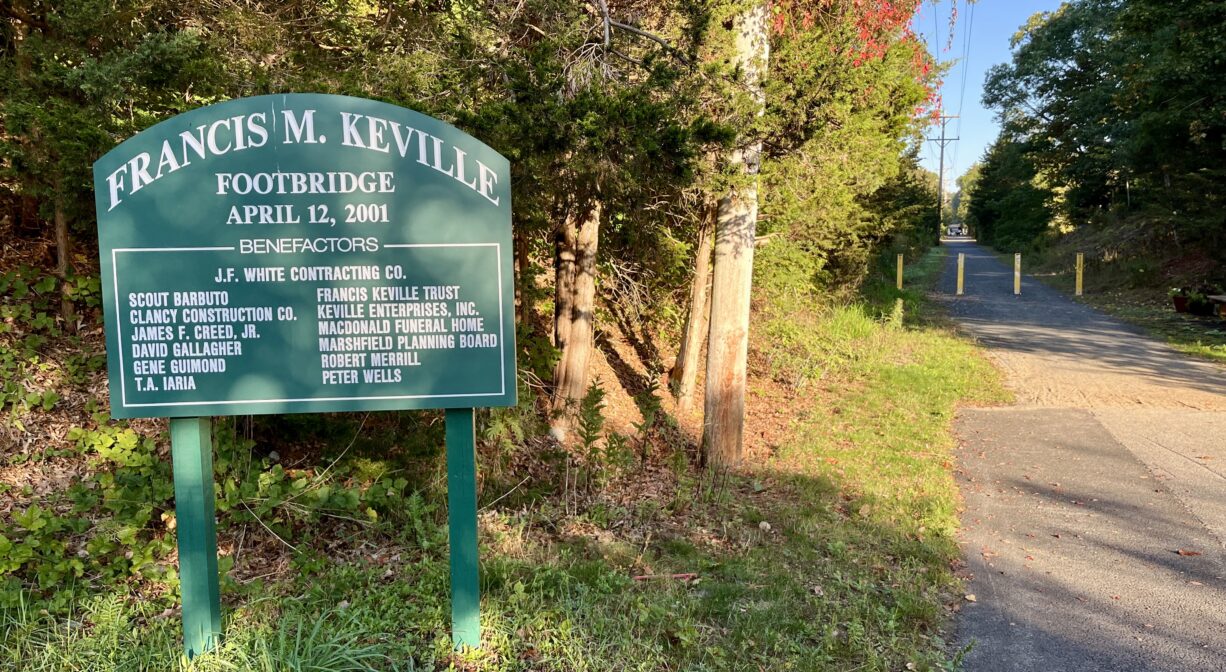 A photograph of a property sign beside a rail trail, with a row of trees in the background.