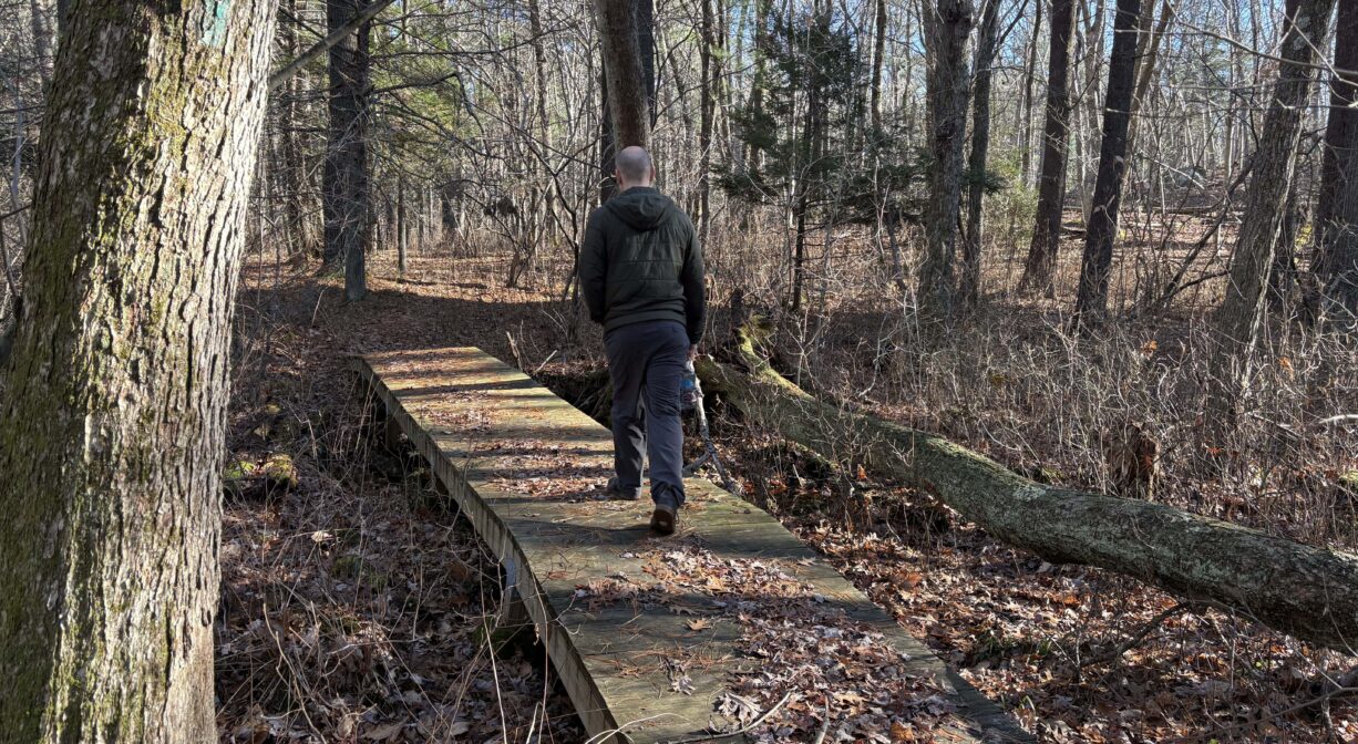 A photograph of an individual walking across a footbridge in a forest.