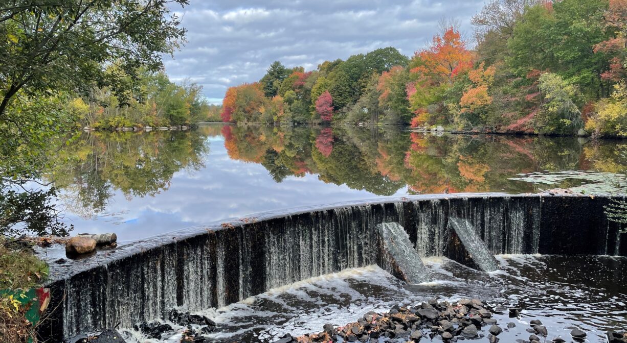 A photograph of a dam with a pond behind it and fall foliage.