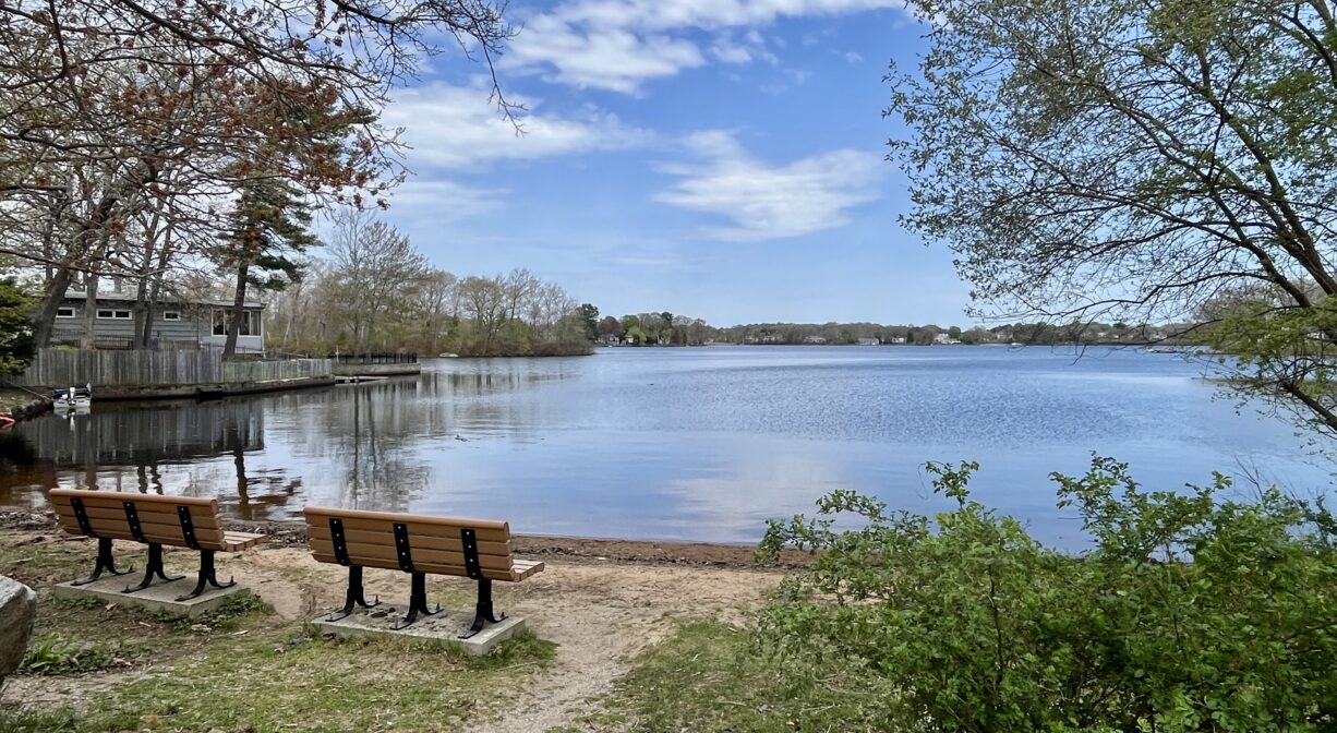 A photograph of two benches on a small sandy beach at a pond.