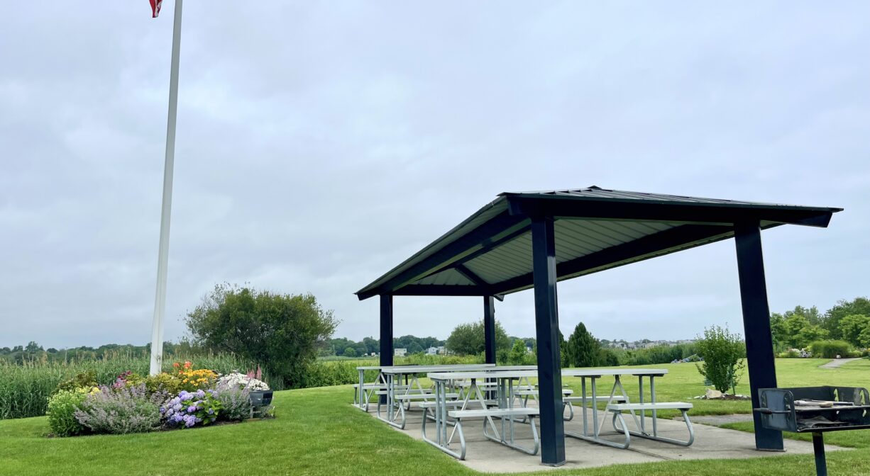 A photograph of a grassy park with a flagpole, a shade pavilion, and several picnic tables.
