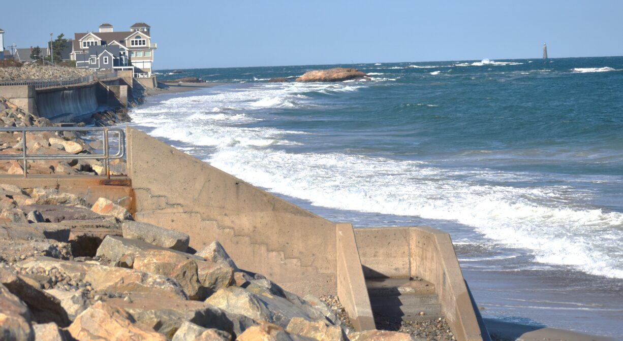 A photograph of an ocean beach with a concrete stairway in the foreground and a lighthouse in the background.