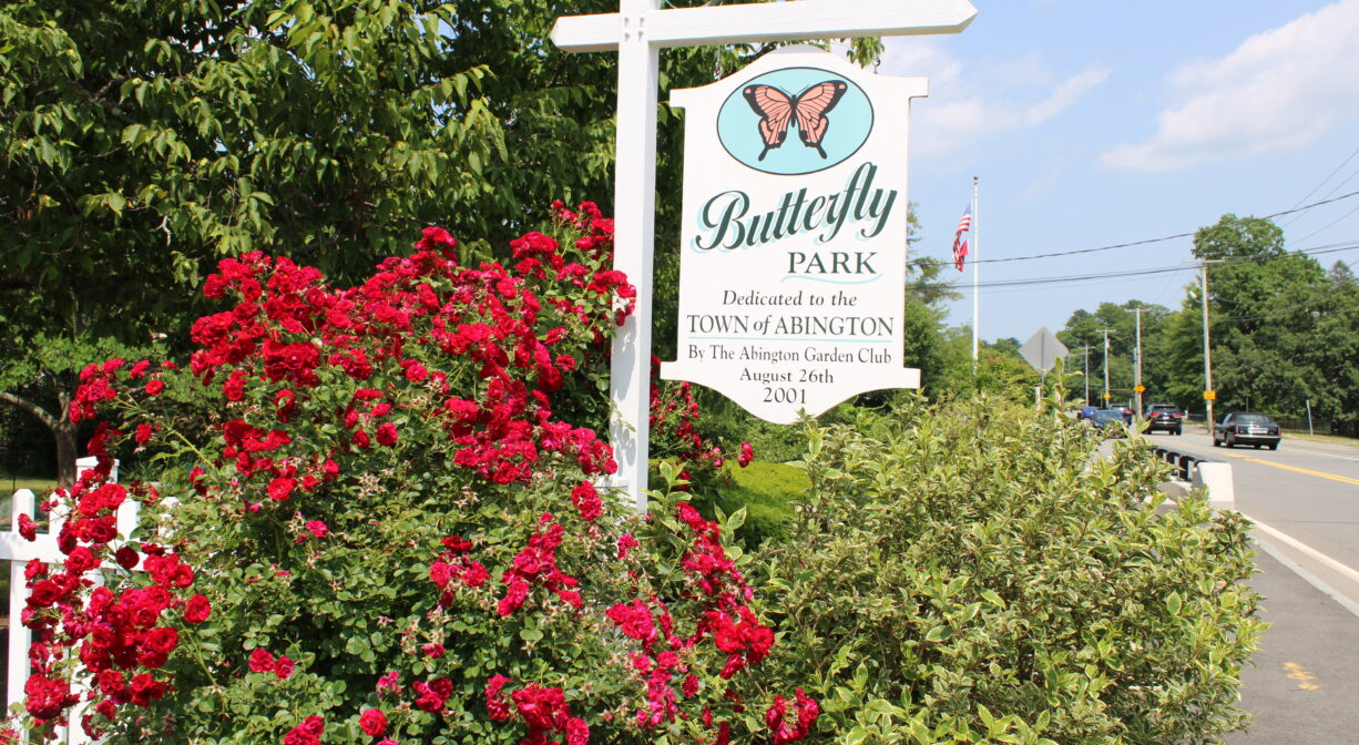A photograph of a property sign surrounded with bushes and red flowers.