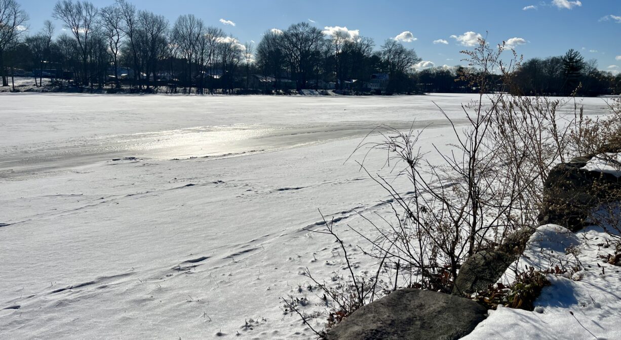 A photograph of a frozen pond, covered with snow, with a cloudy blue sky.