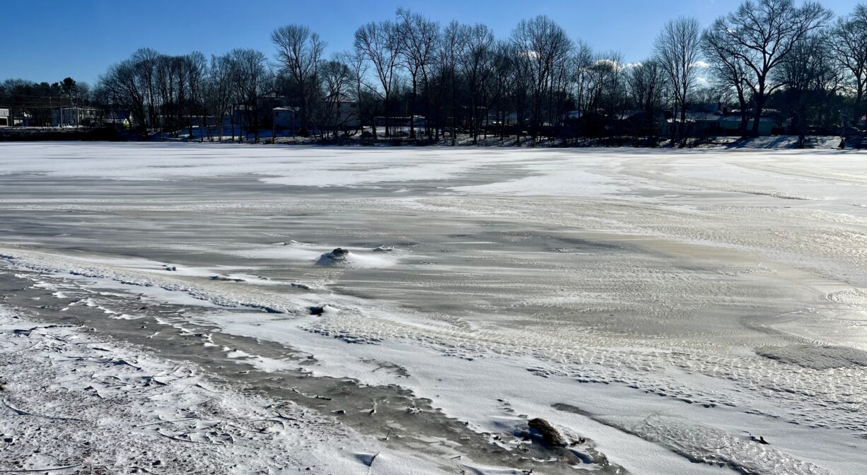 A photograph of a frozen pond, covered with snow, with a cloudy blue sky.