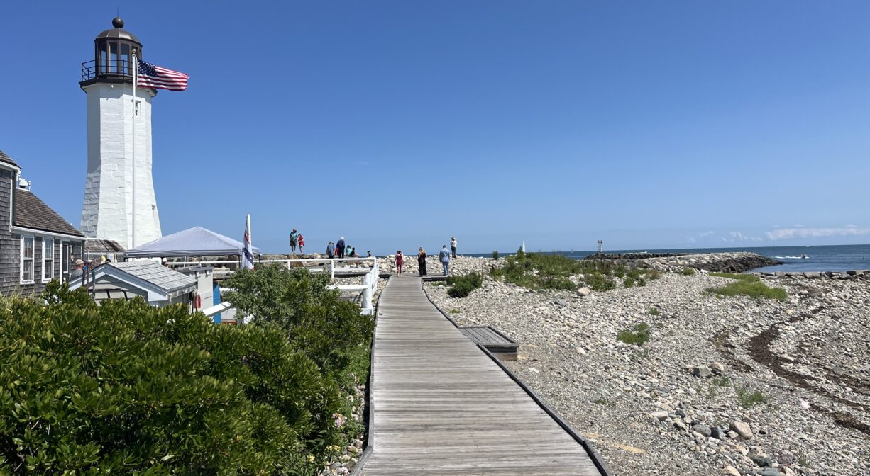 A photograph of a wooden plank walkway leading alongside a beach, to a lighthouse.