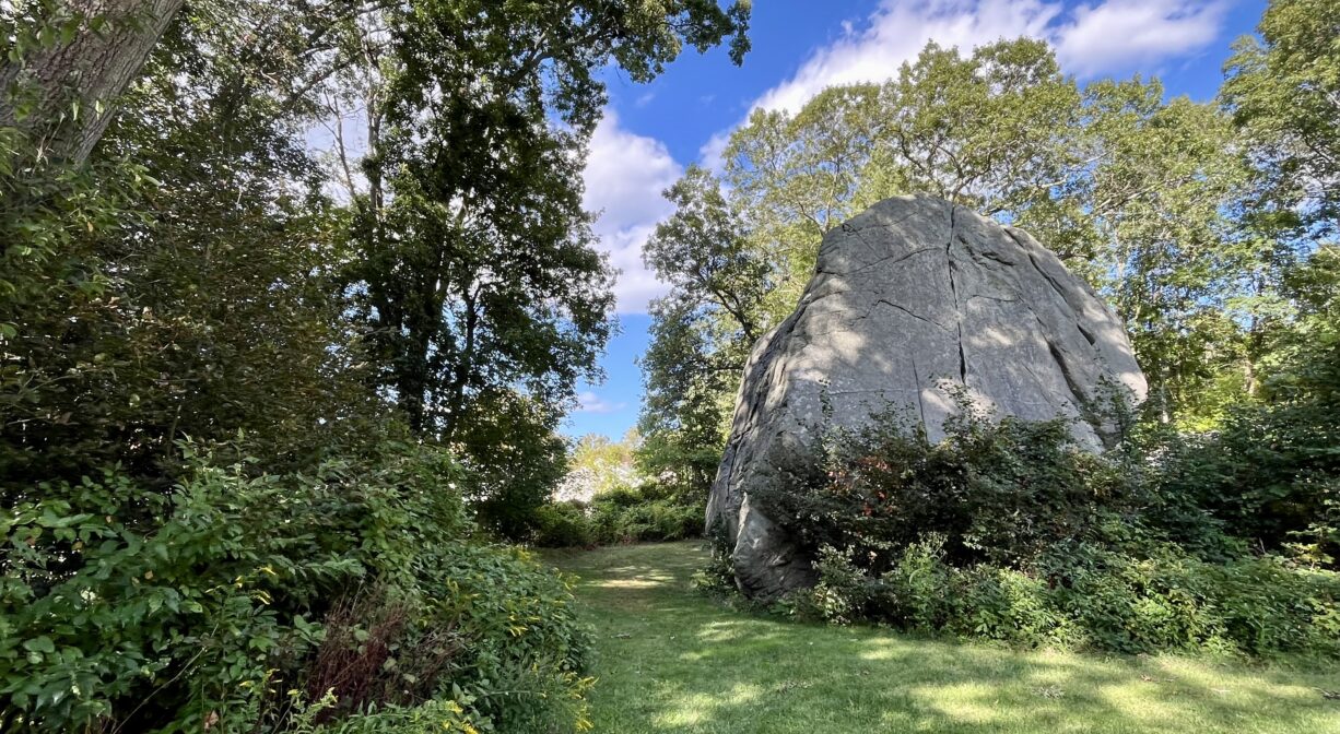 A photograph of a very large boulder within a grassy, tree-lined park.