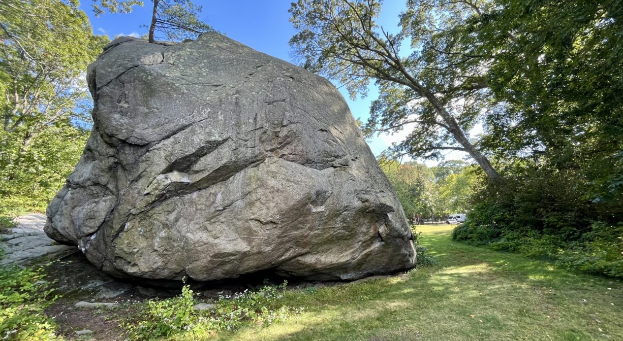 A photograph of a large boulder within a grassy, tree-lined park.