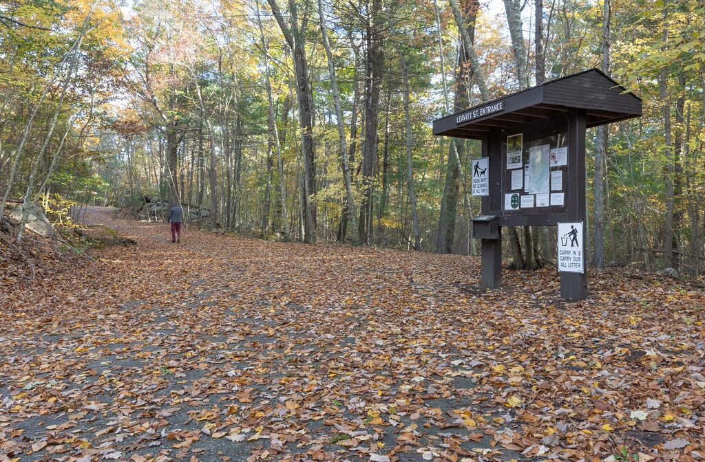 An entrance to a park in the autumn, with fallen leaves and an informational kiosk at a trailhead.