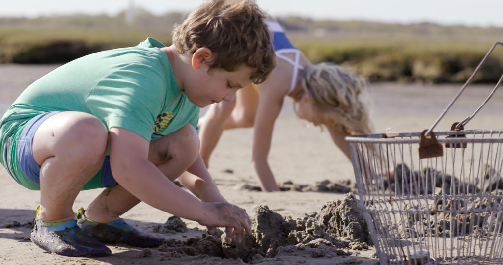 A young boy and a young girl in swim gear digging for clams in the mud at the edge of the salt marsh.