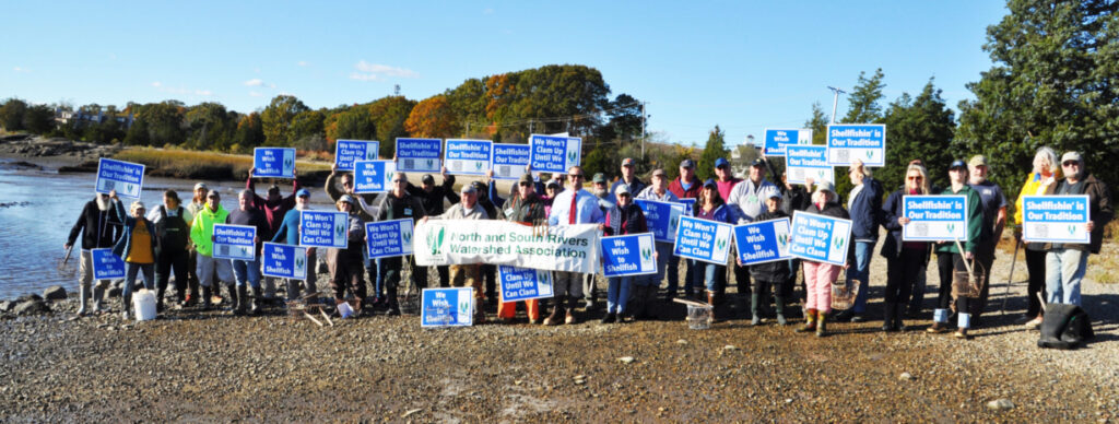 Photograph of a large group of adults and children at the edge of the river with placards.