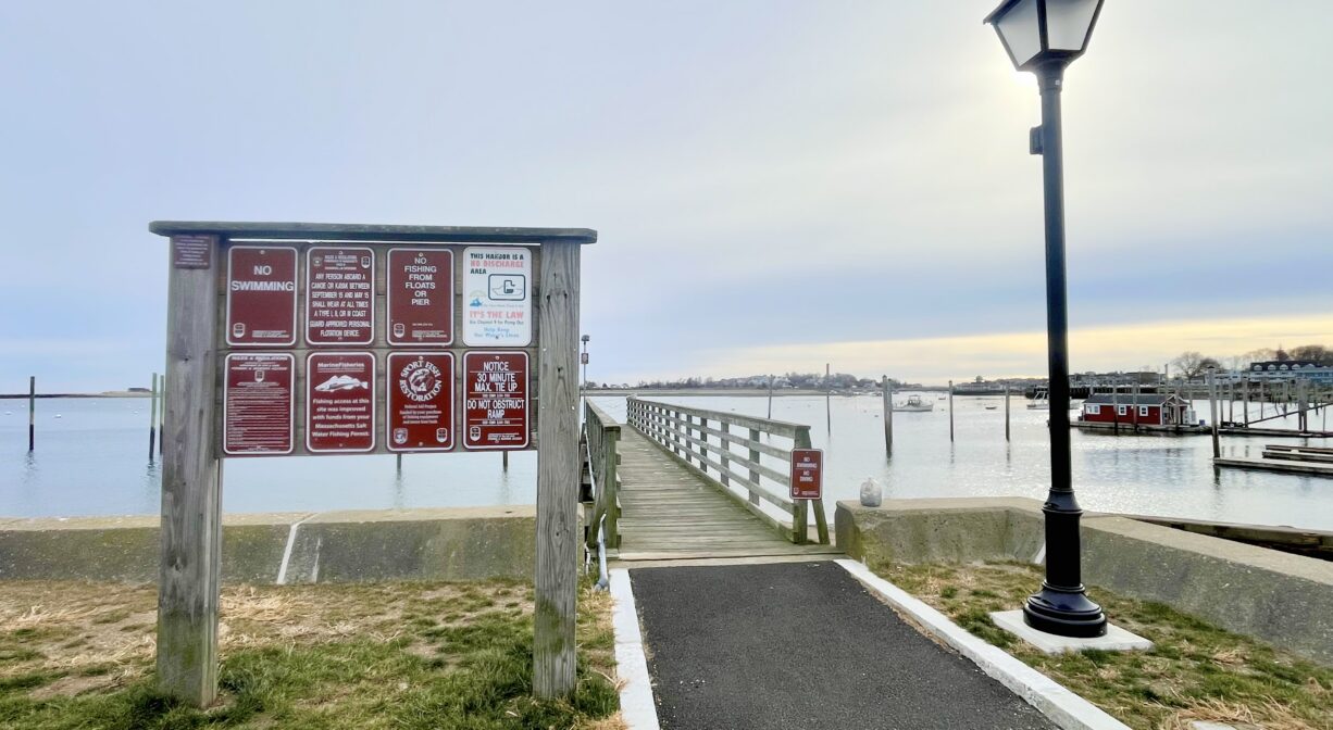 A photograph of a property sign, a paved walkway, and a wooden pier on a harbor.