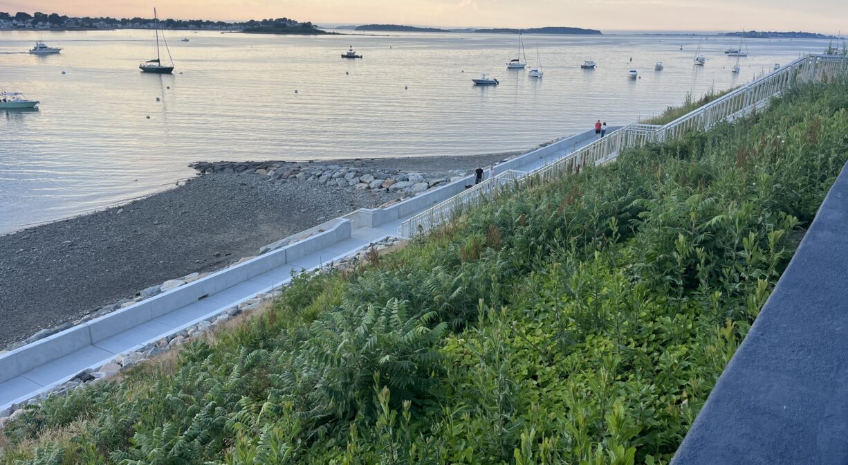 A photograph of a concrete walkway along a shoreline with green vegetation in the foreground.