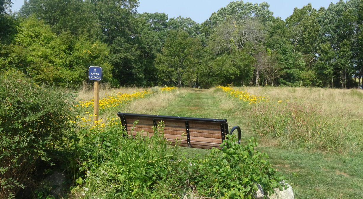 A photograph of a bench in a field with a blue sign to one side.