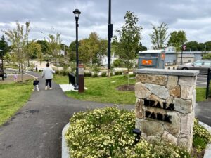 A photograph of a park entrance with paved tails, a property sign, and a mother and child walking in the distance.