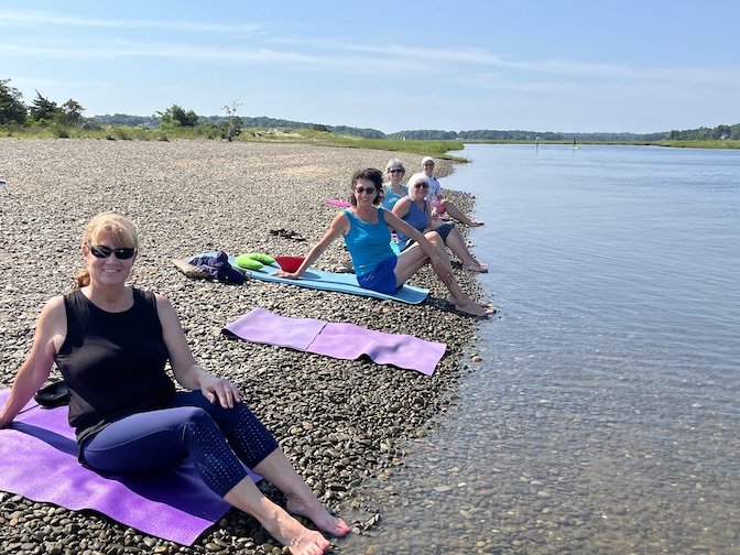 A photograph of a group of women doing yoga directly beside a river.