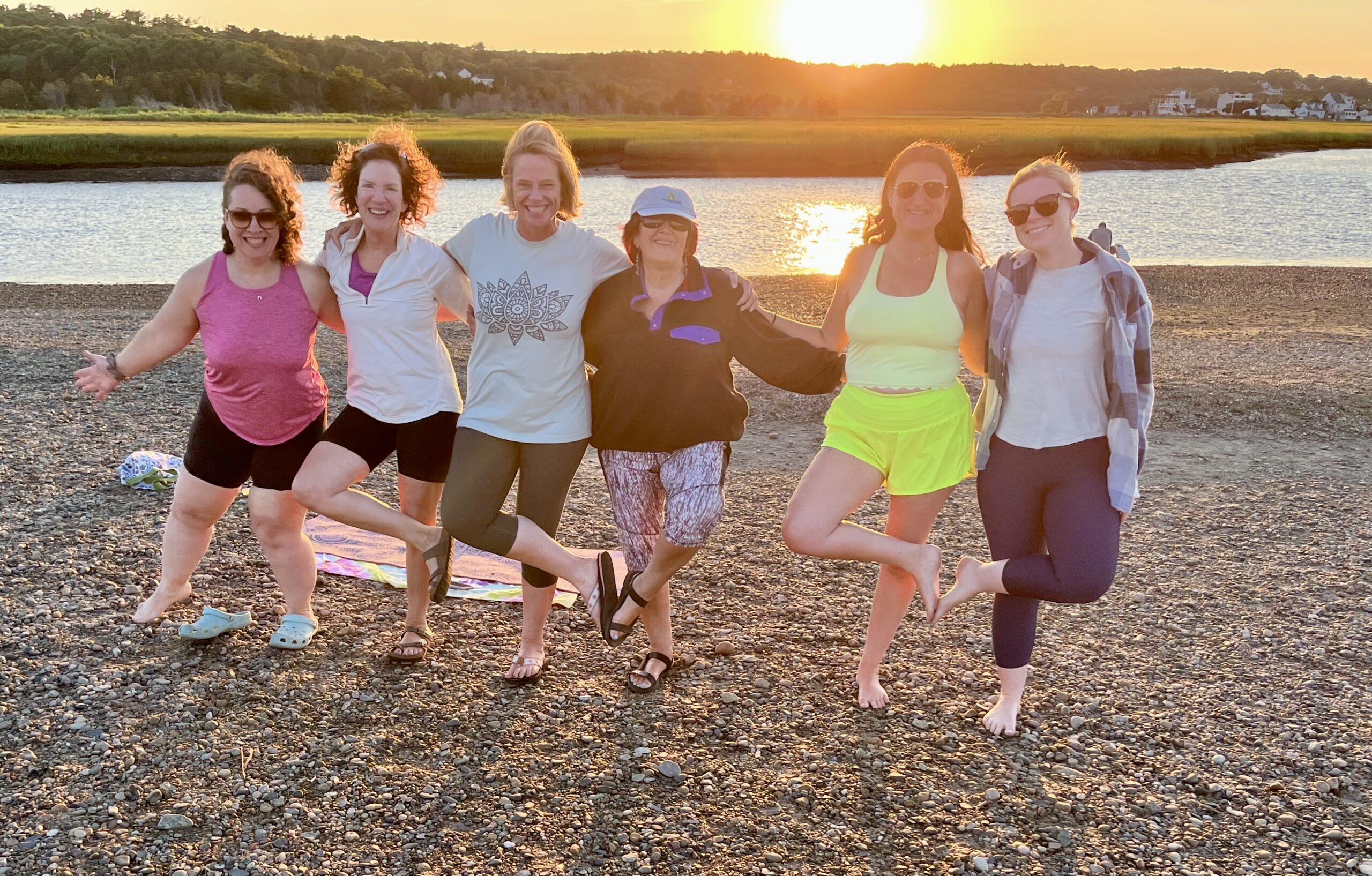 A photograph of a group of women doing the yoga tree pose on a beach by a river.