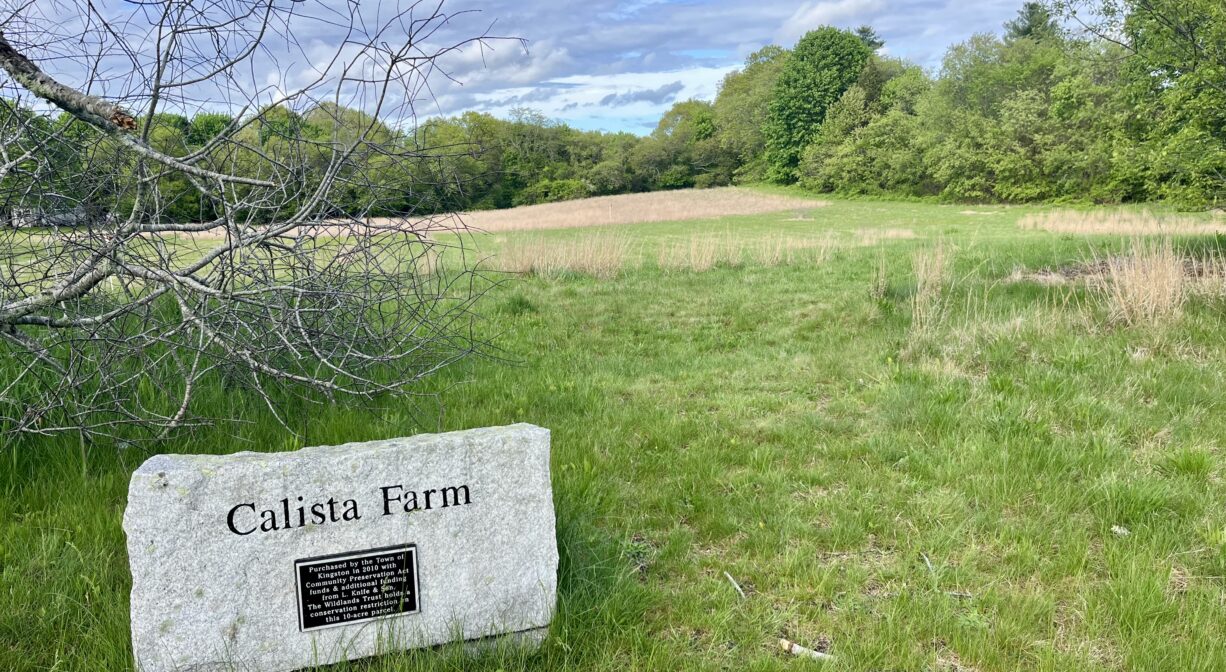 A photograph of a large field with a granite property sign in the foreground.