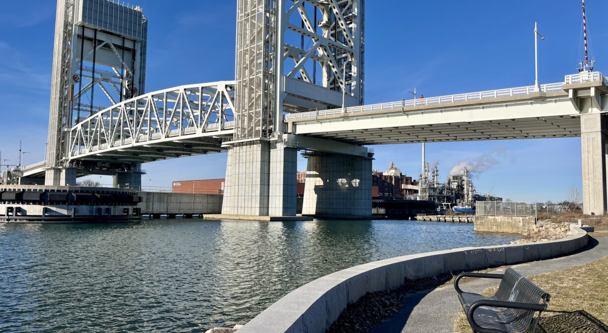 A photograph of a bench overlooking a river, under a large bridge.