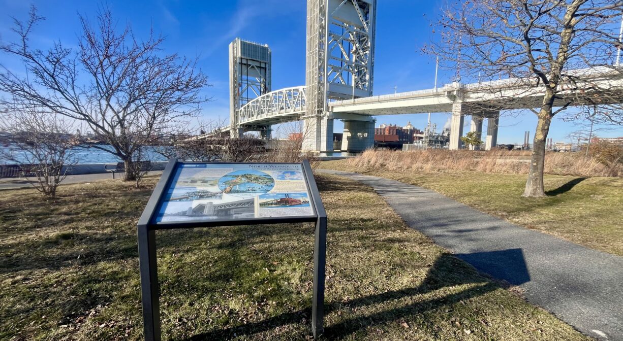 A photograph of an interpretive sign in a park beside a large bridge.