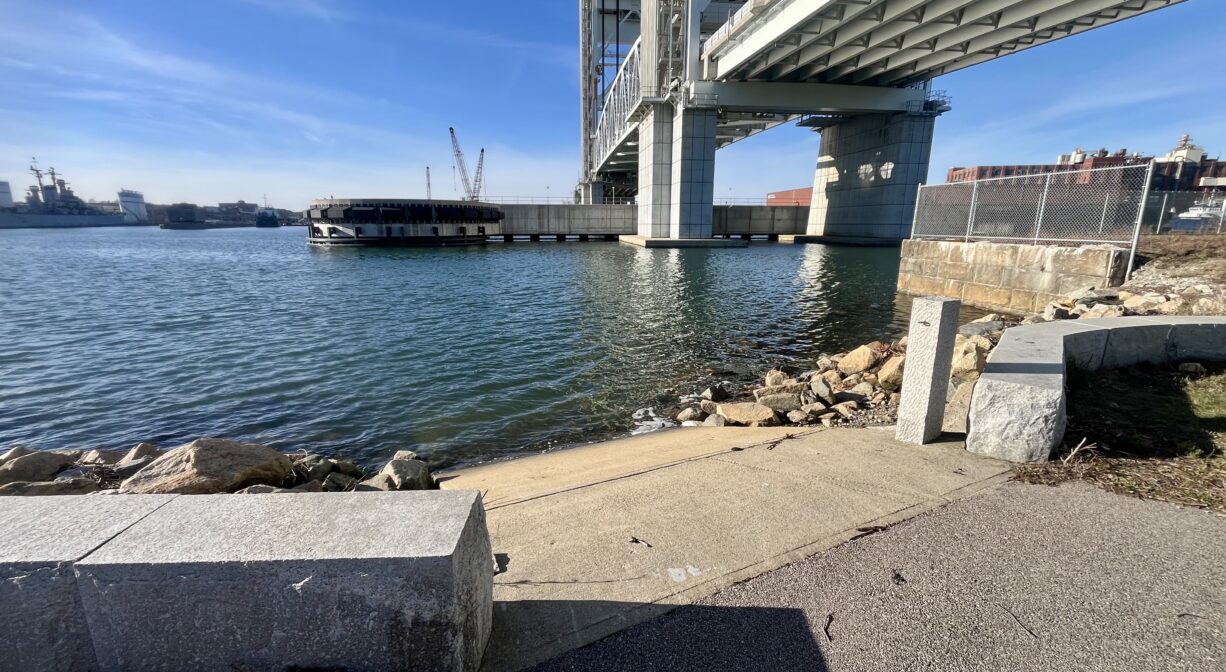 A photograph of a concrete launch area for small boats on a river beneath a bridge.