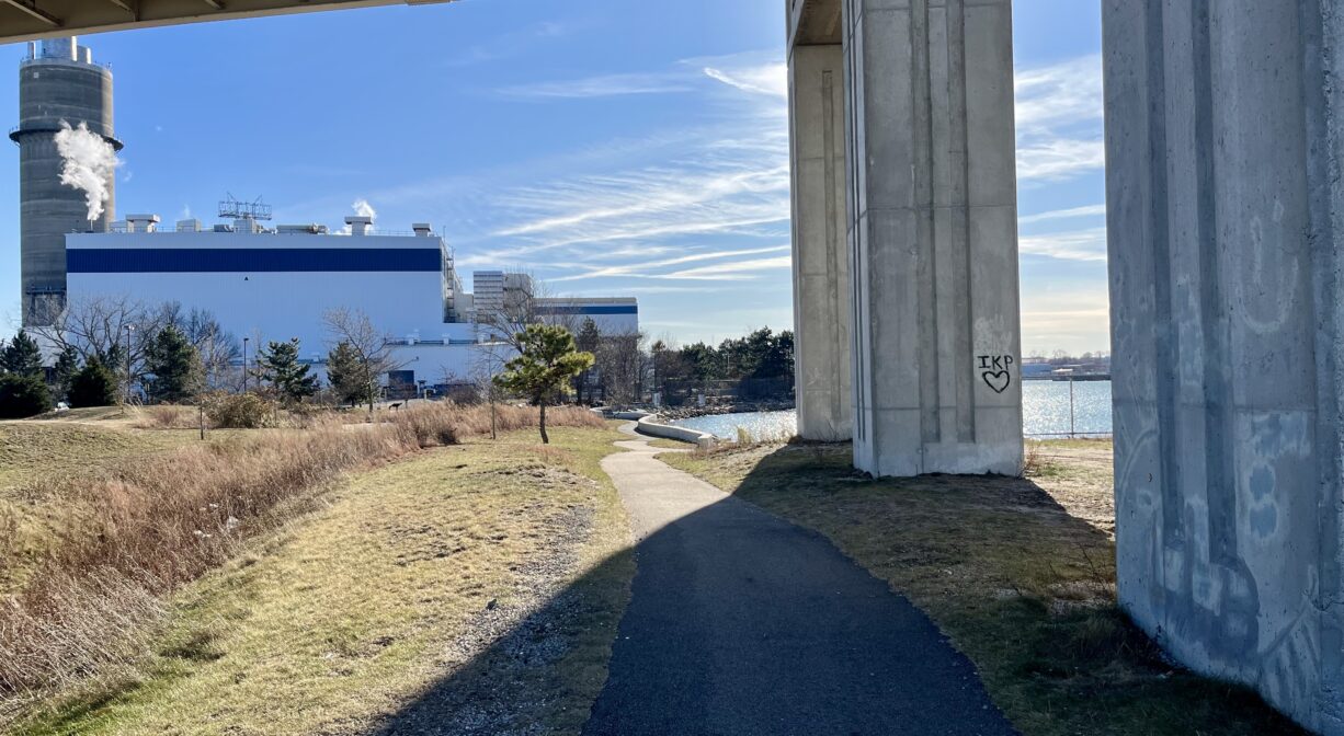 A photograph of a paved trail extending under a bridge by a river.