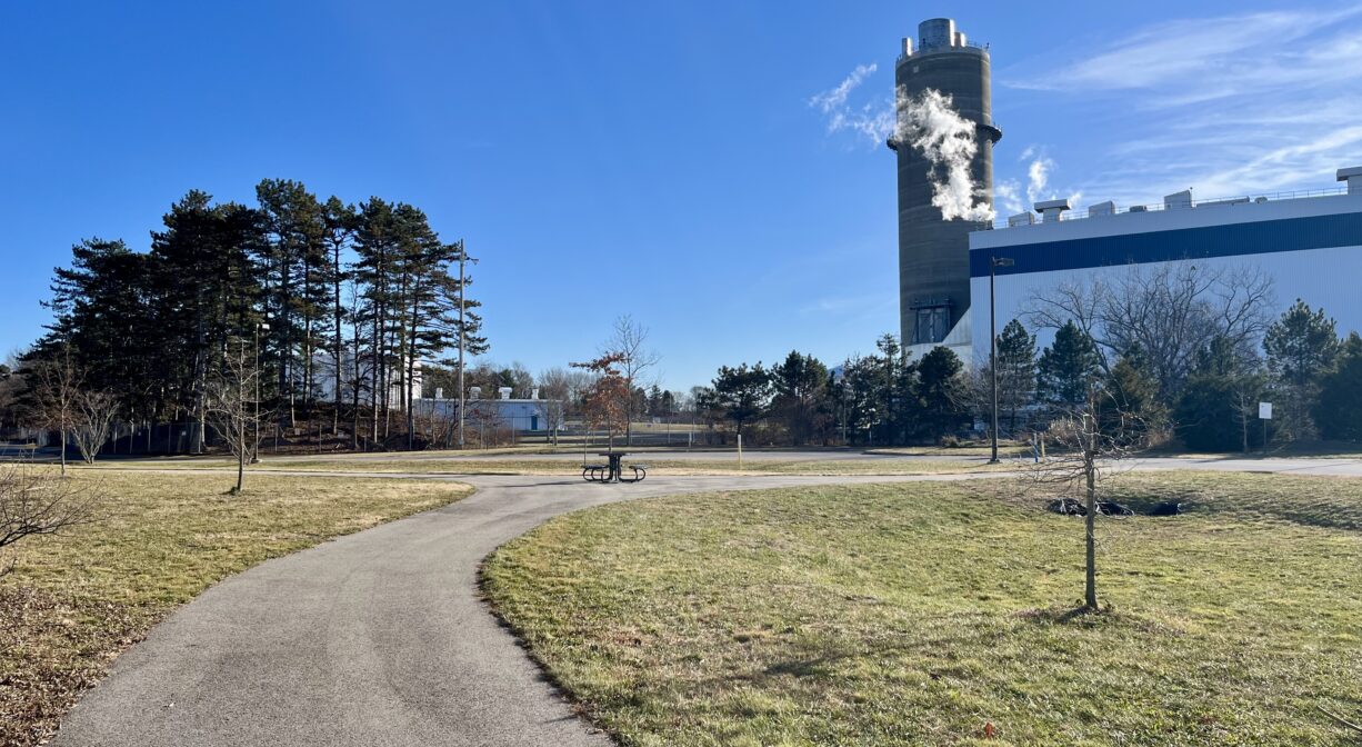 A photograph of a paved trail through a grassy park with a picnic table and an industrial building in the distance.