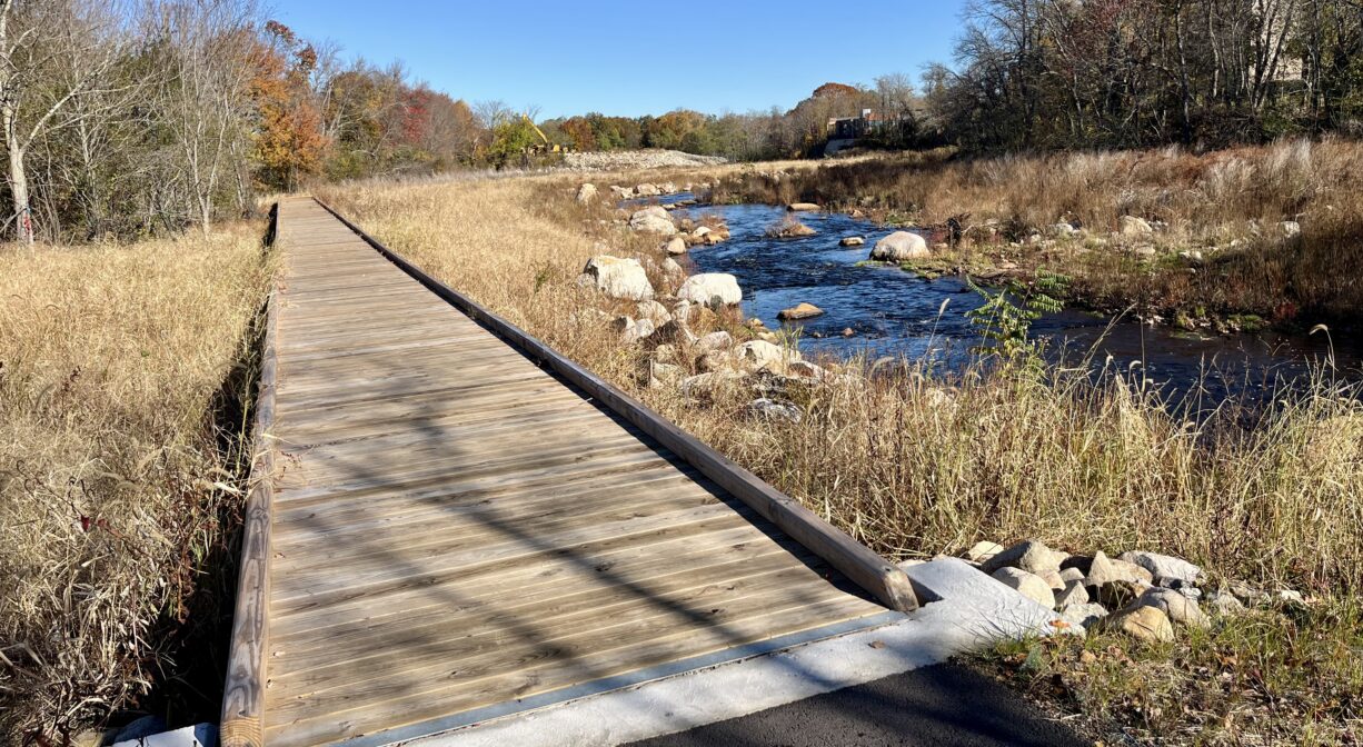 A photograph of a boardwalk extending into a marsh beside a river.