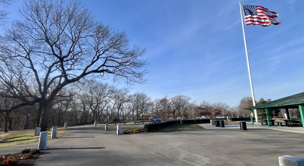 A photograph of a paved plaza in a park with a flagpole and a picnic pavilion.