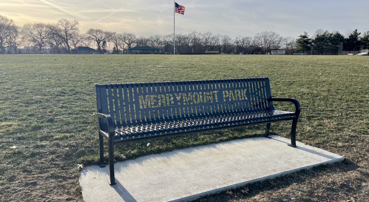 A photograph of a bench in a grassy park, with text on the bench that reads Merrymount Park.