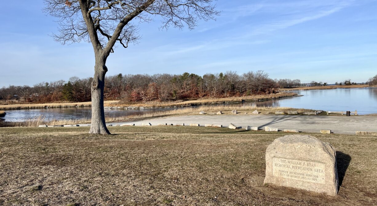 A photograph of a park overlooking a salt marsh with one tree, a property sign, and a large unpaved parking area.