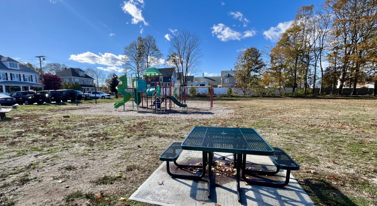 A photograph of a park with a picnic table in the foreground and a playground in the background.
