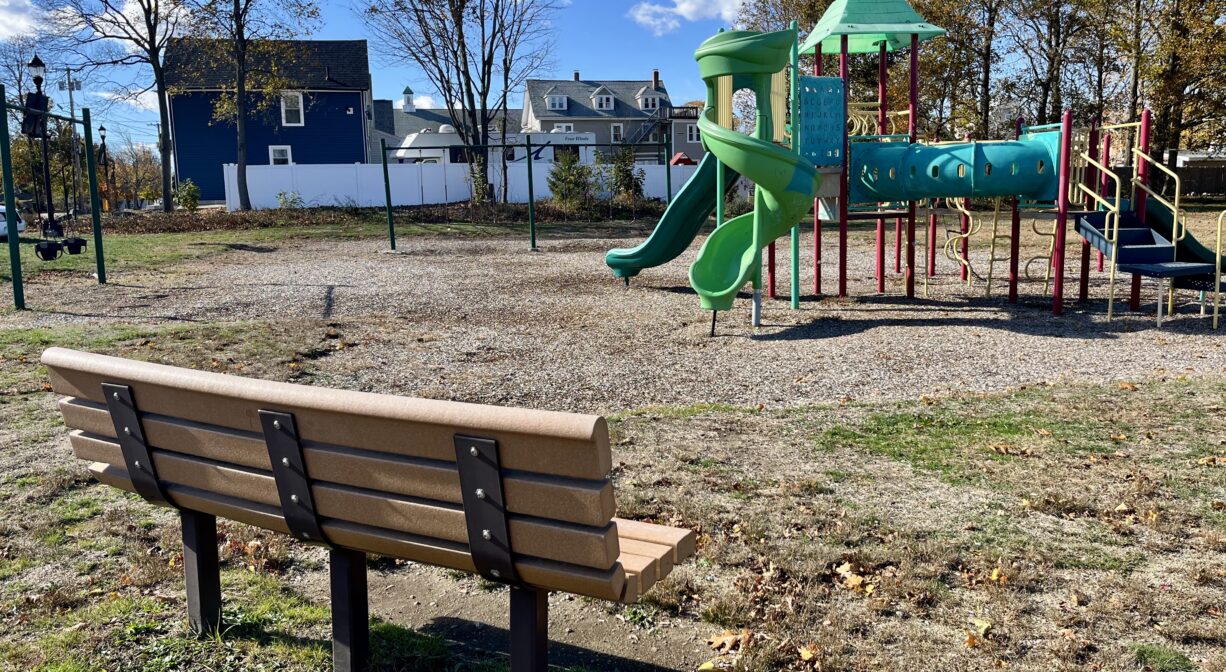 A photograph of a park with a bench in the foreground and a playground in the background.