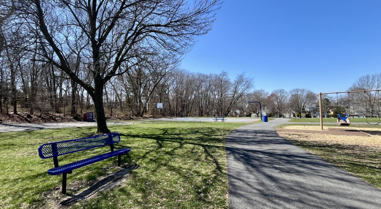 A photograph of a grassy park with a bench and a paved trail.