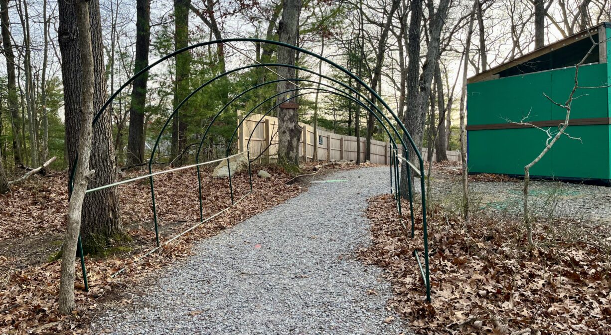 A photograph of a wide crushed stone path through a woodland, with structures to one side.