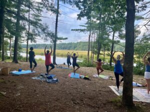 A photograph of a yoga class in a forest by a river.